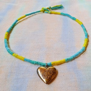 May include: A yellow and blue string necklace with a gold heart-shaped locket. The locket has a floral design.