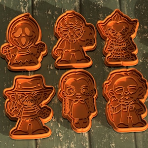 May include: Six orange cookie cutters shaped like horror movie characters. The characters include a ghost, a masked killer, a clown, a girl with pigtails, a man with a hat, and a man with a beard.