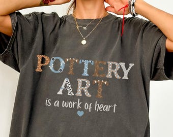 May include: Dark grey t-shirt with the words "POTTERY ART" in a decorative font. The text "is a work of heart" is below the main text, with a small blue heart. The shirt is a Comfort Colors brand.