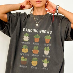May include: Charcoal grey t-shirt featuring the words "DANCING GROWS" above drawings of potted plants, each representing a positive quality such as "Compassion" and "Kindness." The shirt also has the text "Cozy Care" and "Comfort Colors Pepper."