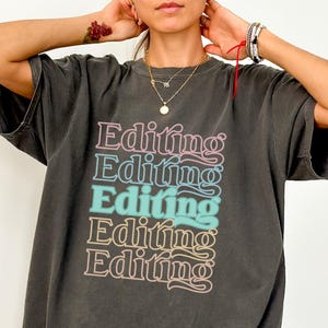 May include: Dark gray t-shirt with the word "Editing" stacked five times in a retro font. The word is in a gradient of pink, blue, and tan colors. The shirt is a Comfort Colors brand.