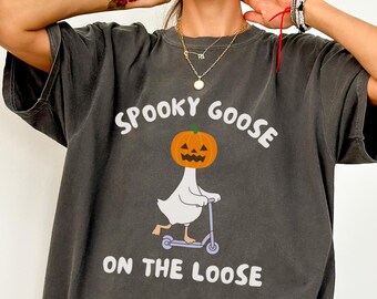 May include: Dark gray t-shirt with the text "Spooky Goose On The Loose". The graphic features a white goose with a pumpkin head riding a scooter. The shirt is a Comfort Colors brand.