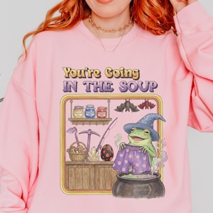 Könnte beinhalten: Hellrosa Sweatshirt mit Rundhalsausschnitt und dem Text "You're Going In The Soup" sowie einer Cartoon-Froschfigur mit Hexenhut und lila Shorts. Der Frosch sitzt in einem Kessel. Das Sweatshirt hat lange Ärmel.