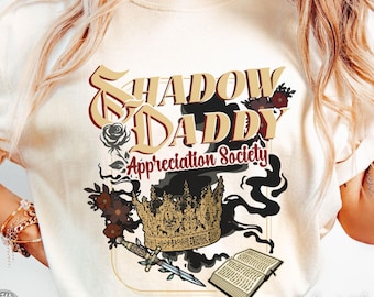 May include: Ivory-colored t-shirt with the text "Shadow Daddy Appreciation Society" in a vintage font. The design includes a gold crown, a dagger, a book, and floral accents. The shirt is a Comfort Colors brand.