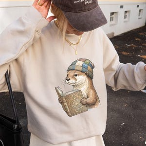 May include: Sand-colored crewneck sweatshirt featuring an illustration of an otter wearing a plaid hat and reading a book. The sweatshirt has long sleeves and a relaxed fit. The text "Sand Crewneck Sweatshirt" is at the bottom.