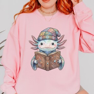 May include: Pale pink crewneck sweatshirt with a colourful illustration of an axolotl wearing a patchwork hat and reading a book. The axolotl is light blue with detailed gills. The text "Cozy Caro" is in the bottom left corner.