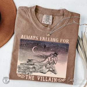 May include: Espresso-coloured Comfort Colors t-shirt featuring the text "ALWAYS FALLING FOR THE VILLAIN" and a graphic of a couple embracing under a crescent moon. A silver necklace with a pendant is draped on the shirt.