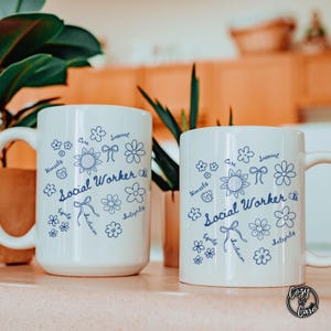 Könnte beinhalten: Zwei weiße Keramikbecher mit blauem Blumendesign und den Worten "Social Worker" in Schreibschrift. Die Becher zeigen auch Wörter wie "Care", "Support", "Honesty", "Equity" und "Integrity".