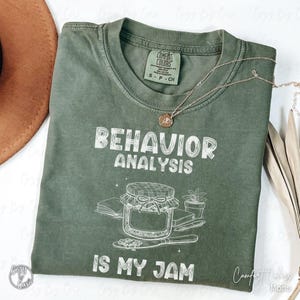 Könnte beinhalten: Ein moosgrünes Comfort Colors T-Shirt mit den weißen Worten "BEHAVIOR ANALYSIS IS MY JAM". Die Grafik zeigt ein Glas Marmelade, Bücher und einen Löffel. Eine goldene Halskette mit Anhänger liegt auf dem Shirt.