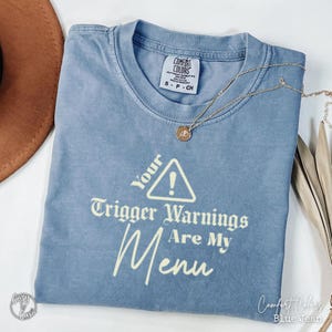 May include: A blue Comfort Colors t-shirt with the text "Your Trigger Warnings Are My Menu". A gold necklace with a pendant rests on the shirt. The shirt is size small. The t-shirt is a blue jean colour.