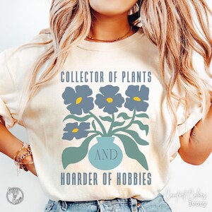 Puede incluir: Una camiseta color marfil con un gráfico de un jarrón azul que contiene tres flores azules con centros amarillos. El texto dice COLLECTOR OF PLANTS AND HOARDER OF HOBBIES en letras mayúsculas azules, con la palabra AND centrada en el jarrón.