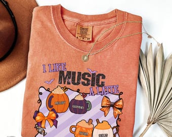 May include: Orange Comfort Colors t-shirt with the phrase "I Like Music A Latte" and pumpkin-themed latte art. The design includes pumpkins, bows, and the words "Melody", "Harmony", "Rhythm", and "Scales". A gold necklace is draped across the shirt.