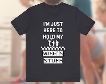 May include: A dark gray t-shirt with the text "I'm just here to hold my wife's stuff" in white. The text is surrounded by a black and white checkered pattern. There are three stick figures running in the checkered pattern.
