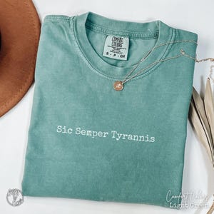 May include: A light green t-shirt with the text "Sic Semper Tyrannis" printed in white. The shirt is made of a soft, comfortable material and has a distressed look.
