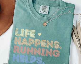 May include: A light green t-shirt with the text "Life Happens. Running Helps." in rainbow colors. The shirt is made of a soft, comfortable material and has a distressed look.