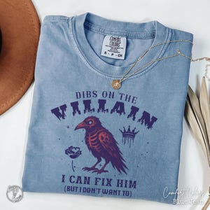 May include: A blue jean Comfort Colors t-shirt with the text "DIBS ON THE VILLAIN I CAN FIX HIM (BUT I DON'T WANT TO)". The shirt features a purple raven illustration with a crown and rose.