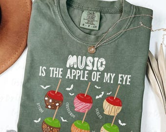 May include: Green Comfort Colors t-shirt with the phrase "MUSIC IS THE APPLE OF MY EYE" in white. The shirt features illustrations of candy apples with musical terms. A gold necklace rests on the shirt.