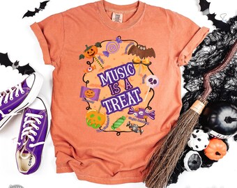 May include: Orange Comfort Colors t-shirt with the text "MUSIC IS A TREAT" surrounded by Halloween-themed graphics. The design includes pumpkins, candy, and a bat. Purple Converse sneakers and a broom are also visible.