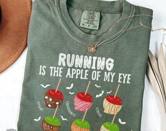 May include: A sage green Comfort Colors t-shirt with the phrase "RUNNING IS THE APPLE OF MY EYE" in white. The shirt features illustrations of candy apples with various toppings and the words "SPRINT", "ENDURANCE", and "STRIDE". A gold necklace rests on the shirt.