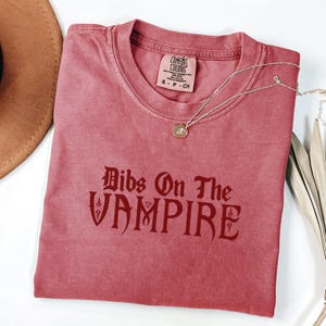 May include: A dusty rose-colored t-shirt with the phrase "Bibs On The Vampire" in a gothic font. The shirt has a crew neck and short sleeves. A silver necklace with a round pendant rests on the shirt. The shirt is folded.