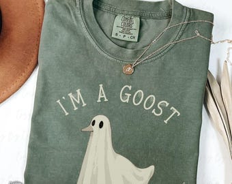 May include: An olive green Comfort Colors t-shirt with the phrase "I'M A GOOST" and a ghost-goose graphic. A gold necklace with a pendant rests on the shirt. The shirt is size S-P-CH.