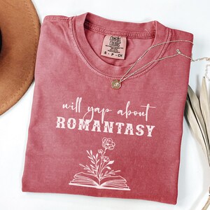 May include: A faded rose-coloured t-shirt with the text "will yap about ROMANTASY" in white. A floral design is below the text. A gold pendant necklace and a brown hat are also visible.