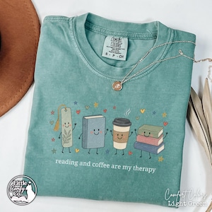May include: A light green t-shirt featuring a graphic of a bookmark, a book, a coffee cup, and a stack of books with faces, arms, and legs. The text reads, "reading and coffee are my therapy," surrounded by small colorful stars and hearts.