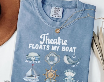 May include: A blue Comfort Colors t-shirt with the phrase "Theatre FLOATS MY BOAT" and theatre-themed graphics. The shirt features a nautical design with a sailboat, life preserver, and whale. A gold necklace is draped across the shirt.