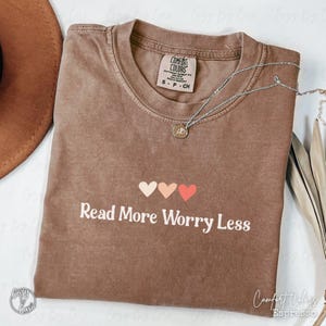 Peut inclure: Un t-shirt marron à manches courtes avec un col rond et une étiquette Comfort Colors à l'intérieur. Le devant présente trois petits cœurs dans des tons crème, pêche et corail au-dessus du texte blanc Read More Worry Less.