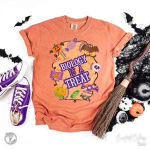 May include: Orange t-shirt with the text "Biology is a Treat" and Halloween graphics. The design includes pumpkins, sweets, a bat, and a skull. Purple Converse trainers are visible on the left.