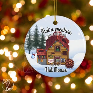 May include: Round Christmas ornament with a winter scene. The ornament features a pixelated cabin with the text "Not a creature was stirring, not even Hat Mouse." A snowman and evergreen trees are also visible.