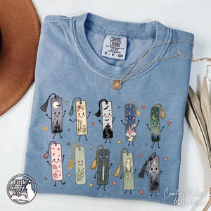 May include: A blue jean colored t-shirt featuring a graphic print of ten anthropomorphic bookmarks with various designs including castles, moons, flowers, and a sword, each with a tassel. The shirt has a Comfort Colors label at the neck.