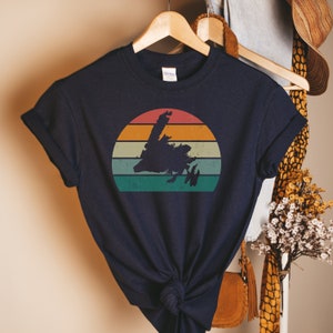 May include: A navy blue t-shirt with a vintage-style sunset graphic featuring the silhouette of the island of Newfoundland. The graphic has a faded, distressed look with shades of orange, yellow, green, and blue.