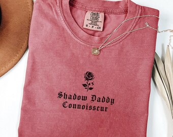 May include: A dusty rose Comfort Colors t-shirt with the words "Shadow Daddy Connoisseur" and a rose graphic. A gold pendant necklace and a brown hat are also visible.