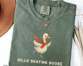 May include: Green Comfort Colors t-shirt with a graphic of a cartoon goose wearing a red scarf and ice skates. The text "SILLY SKATING GOOSE" is below the graphic. A silver necklace with a pendant is on the shirt.