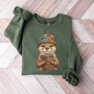 May include: A green sweatshirt featuring a watercolor illustration of an otter wearing a patchwork beanie and holding two books. The otter is brown with a white face and is holding the books with both hands. The books are brown with gold accents.