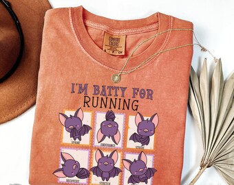 May include: Orange Comfort Colors t-shirt with the phrase "I'M BATTY FOR RUNNING" and cartoon bats on the front. The bats are in different poses with running-related words. A gold necklace and a brown hat are also visible.