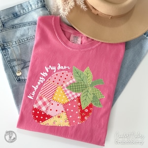 Może przedstawiać: Różowa koszulka Comfort Colors z patchworkowym wzorem truskawki i napisem "Kindness Is My Jam". Truskawka wykonana jest z różnych wzorzystych tkanin. Beżowy kapelusz i niebieskie dżinsy w tle.