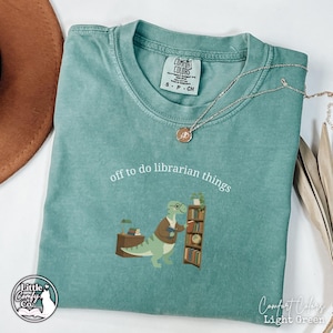 Könnte beinhalten: Ein hellgrünes Kurzarm-T-Shirt mit einer Grafik eines Dinosauriers, der eine Brille und eine Strickjacke trägt und neben einem Bücherregal und einem Schreibtisch steht. Der Text "off to do librarian things" steht in Weiß darüber.