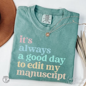 May include: Light green Comfort Colors t-shirt with the text "It's always a good day to edit my manuscript" in pink, blue, and yellow. A gold necklace and a brown hat are also visible.