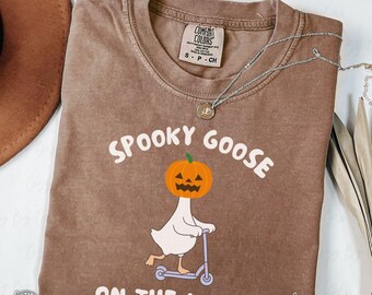 May include: Espresso-colored Comfort Colors t-shirt featuring a Halloween-themed graphic. The design includes a goose with a pumpkin head riding a scooter, with the text "SPOOKY GOOSE ON THE LOOSE". A silver necklace is draped across the shirt.