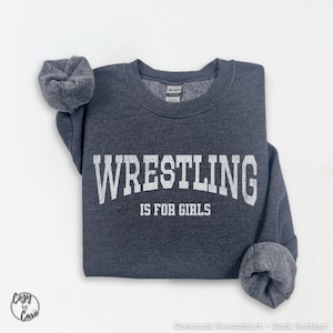 以下が含まれることがあります： 「WRESTLING IS FOR GIRLS」の白文字がプリントされた、濃いヘザーグレーのクルーネックトレーナー。 トレーナーはリブ編みのクルーネックとロールアップされた袖が特徴です。 Gildanブランドのラベルが見えます。