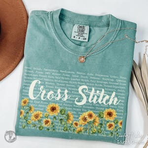 May include: Light green Comfort Colors t-shirt with the words "Cross Stitch" and a sunflower design. The shirt also features a list of cross-stitch related terms. A gold necklace with a pendant is draped on the shirt.