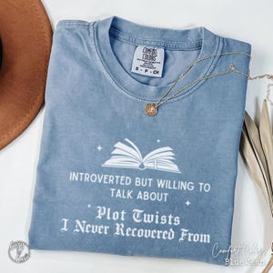 May include: A blue Comfort Colors t-shirt with the text "Introverted but willing to talk about plot twists I never recovered from." The shirt has a white graphic of an open book. A gold necklace with a pendant is on the shirt.