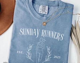 May include: A light blue t-shirt with a white graphic of a woman running. The text "SUNDAY RUNNERS" is above the graphic and "EST. 2025" is below. The shirt is made by Comfort Colors and is in the color Blue Jean.