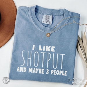 May include: A blue Comfort Colors t-shirt with the text "I LIKE SHOTPUT AND MAYBE 3 PEOPLE" in white. A gold necklace with a pendant is draped across the shirt. The shirt is folded and has a relaxed fit.