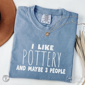 Peut inclure: T-shirt bleu jean Comfort Colors avec le texte blanc "I LIKE POTTERY AND MAYBE 3 PEOPLE". Un collier doré avec un pendentif est posé sur le t-shirt. Un chapeau marron est visible en haut à gauche.