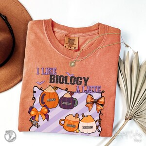 May include: Orange Comfort Colors t-shirt with the phrase "I Like Biology A Latte" and Halloween-themed graphics. The design includes pumpkin-shaped coffee cups and bows, with biology terms like "Mitosis" and "Genetics". A gold necklace and a brown hat are also visible.