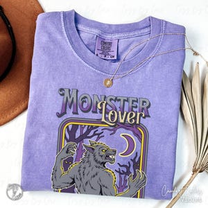 May include: A violet Comfort Colors t-shirt with the text "Monster Lover" in a stylized font. The graphic features a werewolf, a crescent moon, and trees. A gold necklace with a pendant is draped across the shirt.