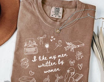 May include: Espresso-colored Comfort Colors t-shirt with the phrase "I like my men written by women" and various line-art illustrations. A silver necklace with a pendant rests on the shirt. The shirt is a size small/ch.